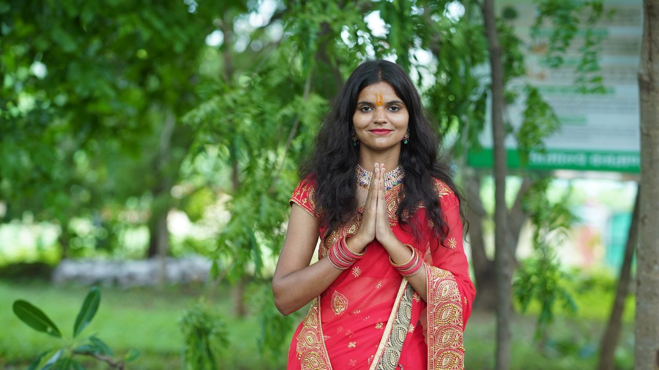 A young woman in a bright red saree standing outdoors, smiling gracefully with hands in a traditional pose.
